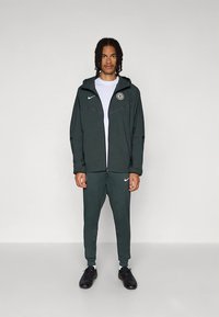 Mørkegrønn Nike-treningsdress med glidelås-hettejakke og avsmalnende bukser, med hvit Nike-logo og en rund logo på venstre bryst.