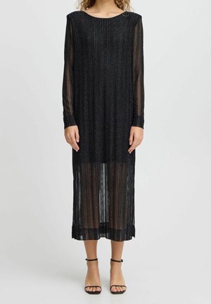 Kobieta w czarnej sukience midi z długim rękawem, ozdobionej pionowymi srebrnymi paskami i przezroczystymi panelami, w połączeniu z czarnymi szpilkami na kostkę z paskiem.