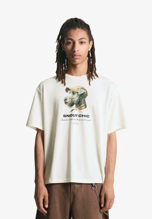 Katoenen oversized crèmekleurig T-shirt met een afbeelding van een hond die een pet draagt en de tekst "SNAOUT CHIC" daaronder. Relaxed fit, korte mouwen.