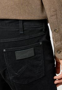 Zwarte corduroy broek met een leren 'Wrangler' label op de achterzak, met contrasterende stiksels en een textuurpatroon.