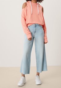 Hoodie corail à épaule dénudée avec cordons blancs, associé à un jean bleu clair à largeur ample et des baskets blanches. Tissu lisse et coupe décontractée.