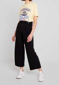 Žluté grafické tričko s motivem UFO a textem, spárováno s černými širokými kalhotami a bílými teniskami. Směs bavlny.