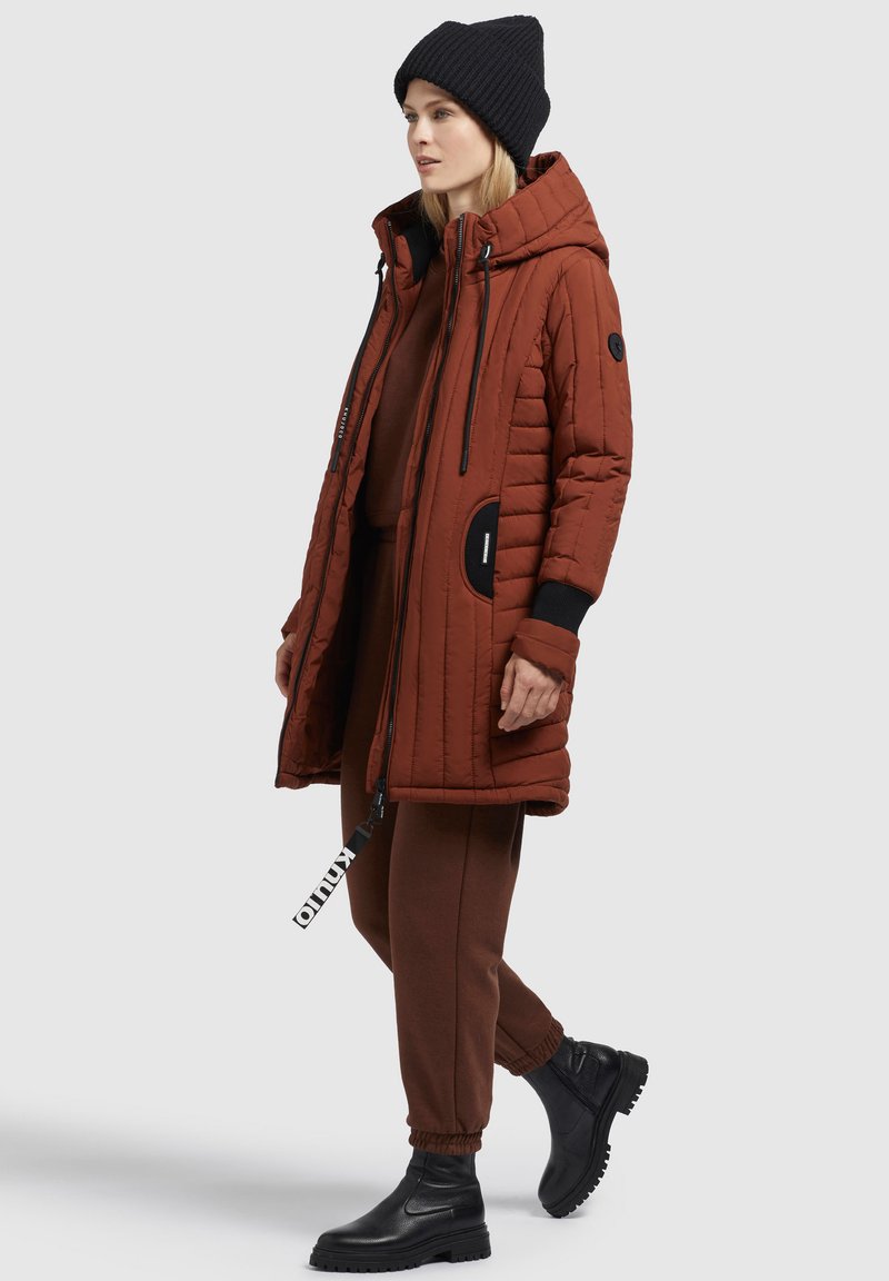 Frau, die einen rostbraunen gesteppte langen Mantel, passende Hose, eine schwarze Strickmütze und schwarze Ankle Boots trägt und vor einem weißen Hintergrund steht.
