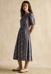 Grün gemustertes Blumenkleid mit kurzen Puffärmeln und gerafftem Taillenbereich. Kombiniert mit weißen Schuhen. Mid-Length-Design.