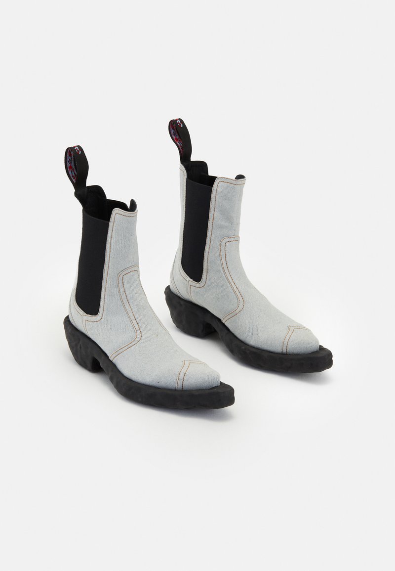 Botas de tobillo de tela gris claro con paneles elásticos negros a los lados, punteras puntiagudas y suelas de goma negra texturizada. Presentan tiradores coloridos.