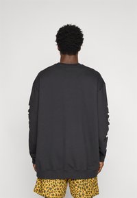 Juodas džemperis su ilgomis rankovėmis, ribbintais rankogaliais ir apačia, su baltu užrašu ant kairės rankovės. Dėvimas su geltonais šortais su leopardo raštu.