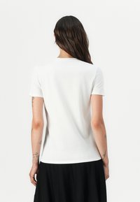 T-shirt ajustée blanche à manches courtes, col rond, fabriquée en tissu lisse, présentée de dos avec une jupe plissée noire.
