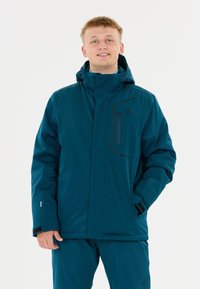 Junger Mann in einer tealfarbenen Winterjacke mit Kapuze und passenden Hosen, lächelnd mit einer Hand in der Tasche vor einem weißen Hintergrund.