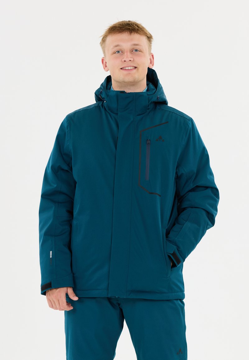 Junger Mann in einer tealfarbenen Winterjacke mit Kapuze und passenden Hosen, lächelnd mit einer Hand in der Tasche vor einem weißen Hintergrund.