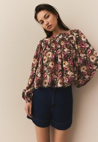 Bluză florală cu mâneci posomorâte lungi, de culoare verde închis, cu flori roz și galbene, asortată cu pantaloni scurți din denim închis.