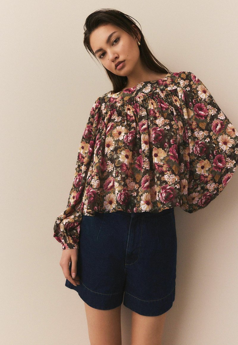 Bluză florală cu mâneci posomorâte lungi, de culoare verde închis, cu flori roz și galbene, asortată cu pantaloni scurți din denim închis.