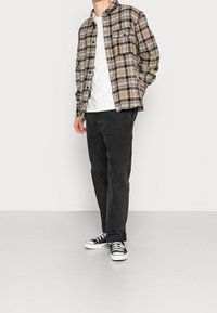 Rutig flanellskjorta i beige, svart och brunt, bärs över en vit t-shirt; ihopparad med svarta byxor och svart-vita sneakers.