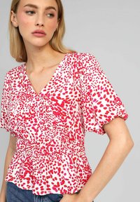 Blouse blanche avec un motif de points abstraits rouges et roses, décolleté en V, manches courtes bouffantes et détail de taille froncé. Convient pour un usage décontracté.