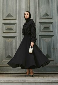 Vestido negro largo con volantes en capas, corsé ajustado y falda acampanada. Accesorios como un pequeño bolso de perlas y zapatos de tacón.