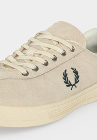 Baskets en daim beige avec lacets crème, œillets métalliques, semelle texturée et logo couronne de laurier brodé vert foncé sur le côté.
