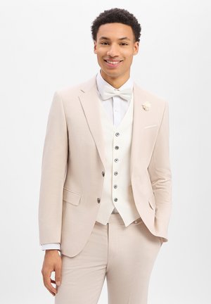 Junger Mann lächelt, trägt einen hellbeigen Anzug mit weißer Fliege, Weste und einer Blumenanstecknadel, steht vor einem schlichten weißen Hintergrund.