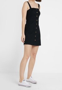 Eine Frau trägt ein ärmelloses schwarzes Kleid mit Knopfleiste und weiße Sneaker. Sie steht vor einem schlichten weißen Hintergrund.