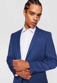 Marineblauwe colbert met notch lapels, zijzakken en een enkele knoopsluiting, over een wit shirt met blauwe verticale strepen.