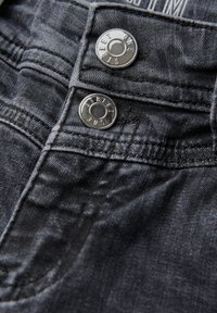 Les jeans en denim foncé présentent deux boutons métalliques avec un marquage gravé. Le tissu a une texture lisse avec des dégradés subtils et des coutures visibles.