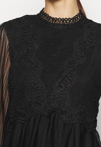 Abito in pizzo nero con collo alto, intricate trame in pizzo e maniche trasparenti nere per un tocco di texture e dettaglio.