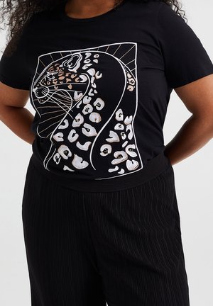 Czarny t-shirt z haftowaną stylizowaną głową lamparta w białym, beżowym i złotym kolorze, noszony z czarnymi plisowanymi spodniami, ręce za plecami.