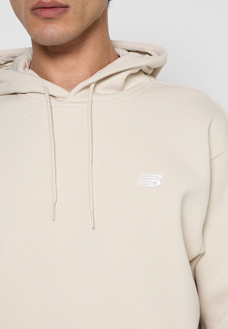 Mann trägt beige Hoodie mit Kordeln und kleinem weißem New Balance-Logo auf der Brust, vor einem einfarbigen hellen Hintergrund.