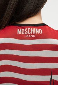 Widok z tyłu osoby z długimi ciemnymi włosami, ubraną w czerwoną i białą bluzkę w paski z napisem "MOSCHINO JEANS" na górnej części pleców.