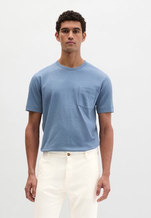 Giovane uomo con capelli ricci che indossa una maglietta a maniche corte azzurro chiaro con taschino sul petto e pantaloni panna, in piedi contro uno sfondo bianco uniforme.