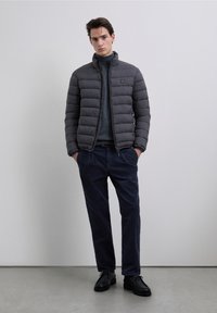 Szara kurtka puchowa z wysokim kołnierzem i przeszywanym wzorem, zestawiona z ciemnym golfem, granatowymi spodniami z kordurowej tkaniny oraz czarnymi butami.