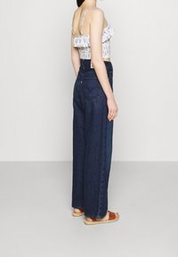 Bílý květinový crop top s volány je kombinován s vysoce pasovými, volnými tmavě modrými džínami. Jsou nošeny sandály s tkanou podrážkou.