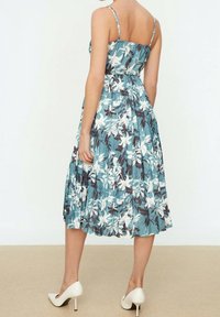 Eine Frau trägt ein ärmelloses Midikleid mit blauen und weißen Blumen und Falten. Sie steht barfuß auf einem beigen Boden vor einer einfarbigen hellen Wand.