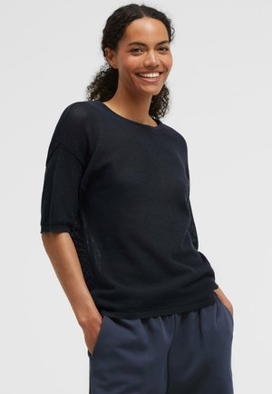 Smiling woman with curly hair wears a black knit half-sleeve top and dark pants, standing with hands in pockets against a plain background.