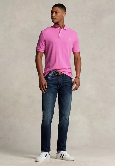 Un giovane si erge con sicurezza indossando una polo rosa e jeans scuri, con scarpe da ginnastica bianche e una cintura a righe blu, su uno sfondo neutro.