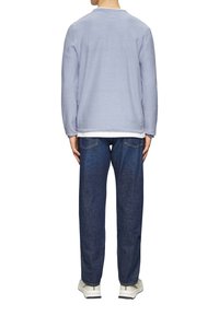 Maglione di cotone azzurro chiaro con colletto tondo, indossato sopra una camicia bianca, abbinato a jeans in denim blu scuro e sneaker beige.