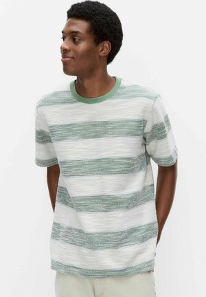 Jonge man met kort krullend haar die een wit-groen shirt met brede strepen en korte mouwen draagt, kijkt naar links met de handen achter de rug.