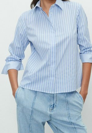 Femme portant une chemise à rayures verticales bleu clair et blanc avec manches retroussées et un jean en denim bleu clair, les mains dans les poches.