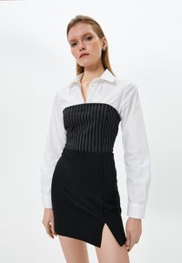 Abito bianco e nero con un corpetto a righe strette aderente e una gonna nera corta con uno spacco laterale. Realizzato in tessuto morbido con colletto.