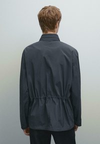 Giubbotto a vento grigio scuro con vita arricciata e colletto eretto. Realizzato in materiale leggero con maniche lunghe e una texture liscia.