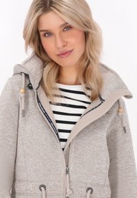 Gai bēšas krāsas zip-up jakas ar teksturētu apdari, ar lielu kapuci, auklām un jūras zila krāsas odere apkaklei. Uzvilkta virs svītrainas krekla.