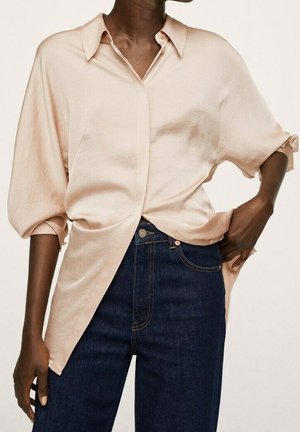 Donkere persoon die een losse lichtroze blouse met knopen en opgerolde mouwen draagt en donkerblauwe, hooggetailleerde spijkerbroek. Hand op de heup.