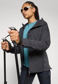 Schwarze Outdoor-Jacke mit strukturierten Einsätzen, getragen über einem petrolfarbenen Oberteil. Dunkle Leggings, Trekkingstöcke mit grauen Griffen und eine Sonnenbrille vervollständigen den Look.