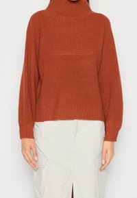 Sweter w kolorze rdzawym, dziergany, z wysokim kołnierzem i luźnym krojem, o fakturze prążkowanej. Zestawiony z jasną spódnicą do kolan.