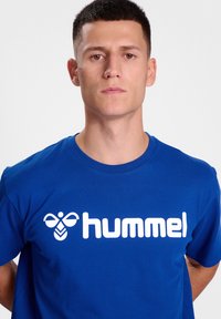 Blå bomull T-shirt med en stor vit "hummel"-logotyp och grafik ovanför, med klassisk rund hals och korta ärmar.