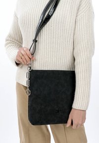 Femme portant un pantalon beige et un pull en maille blanc tenant un sac bandoulière noir texturé avec une chaîne argentée et un charm logo.