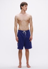 Ανδρικό μπλε ναυτικό μαγιό κοντό κατασκευασμένο από ελαφρύ ύφασμα, με λευκό κορδόνι, πλαϊνές τσέπες και λευκές ραφές για διακόσμηση.