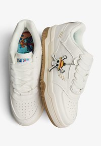 Scarpa da ginnastica bianca con superficie testurizzata, punta tonda e design con lacci. Presenta una suola marrone dorato e un ciondolo con teschio e tibie incrociate.