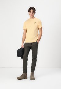 Yellow short-sleeve t-shirt with a small green butterfly logo, paired with olive green pants and beige ankle boots. Holds a black bag.
