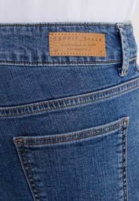 Vaqueros de mezclilla azul con un parche de cuero marrón cosido etiquetado como "ESPRIT Denim" encima del bolsillo trasero y del pasador del cinturón.