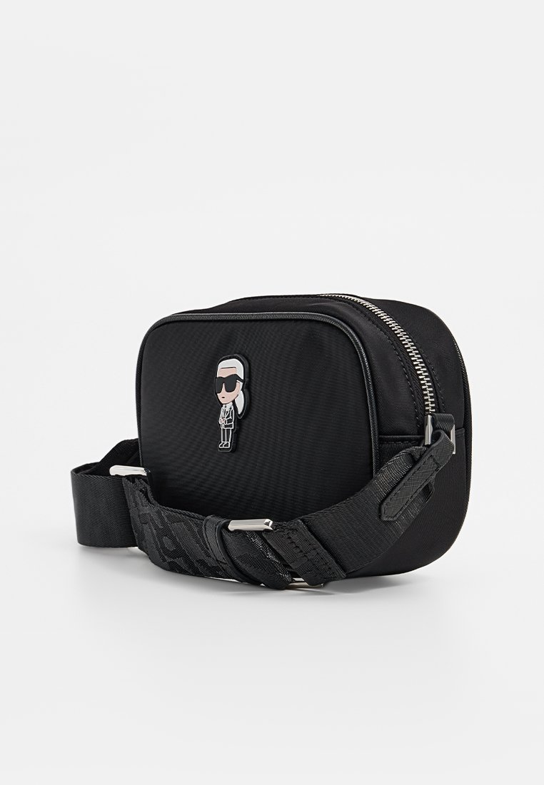Bolso cruzado rectangular negro con correa ajustable, cremallera plateada y pequeño emblema de un personaje con gafas de sol en la parte frontal.
