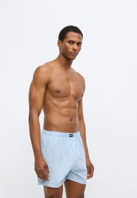 Ανοιχτό μπλε μαγιό σορτς με ελαστική μέση, κατασκευασμένο από απαλό ύφασμα. Διαθέτει μαύρη ετικέτα μάρκας στο μπροστινό μέρος.